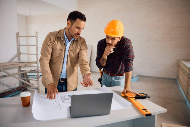 Construction worker and his client using laptop while examining housing plans.